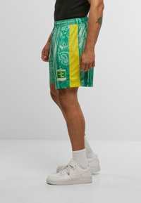 Grønne mønstrede sports-shorts har en gul sidepanel, elastisk talje og et logo-patch. Båret med hvide sneakers og strømper.