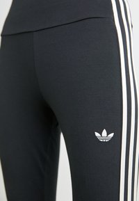Kratki prikaz crnih sportskih tajica s bijelim trostranim bočnim prugama i malim bijelim Adidas Trefoil logom na bedru.