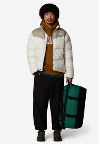 En pufferjacka i krämfärg och olivgrön, ihop med en brun hoodie och svarta byxor. Håller en grön och svart duffelväska. Svarta skor.
