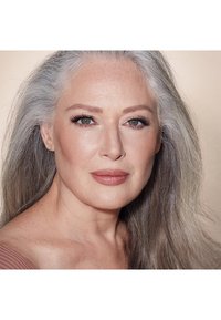Primo piano di una donna con lunghi capelli grigi e lisci, pelle liscia, trucco naturale e colore delle labbra neutro su uno sfondo morbido e neutro.