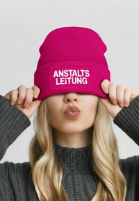 Pink knit beanie with a contrasting rectangular patch printed with "ANSTALTSLEITUNG." Model wears a dark gray textured sweater.