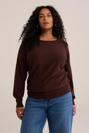 Femme aux cheveux noirs bouclés portant un pull marron à manches longues et un jean bleu, debout devant un fond beige uni.
