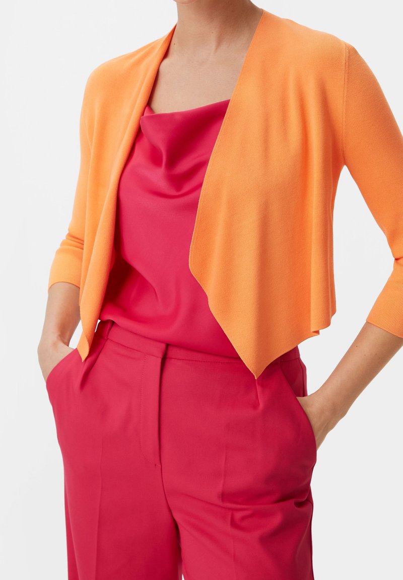 Femme portant un cardigan ouvert orange vif sur un haut rouge sans manches et un pantalon taille haute rouge, les mains dans les poches.