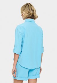 Camicetta azzurro chiaro a maniche corte con risvolti, realizzata in un tessuto testurizzato, caratterizzata da una vestibilità relax e un orlo curvo. Indossata con pantaloni corti abbinati.