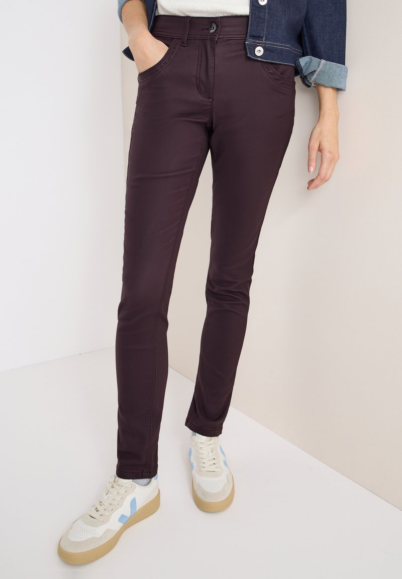 Burgundské slim fit džíny s pěti kapsami, vyrobené z elastického materiálu. V kombinaci s bílými teniskami s modrými akcenty a béžovou podrážkou.
