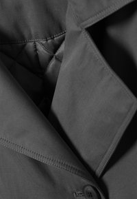Tessuto del giubbotto grigio con una texture liscia. Presenta una fodera interna trapuntata e un dettaglio con bottone. Linee pulite e design minimalista.