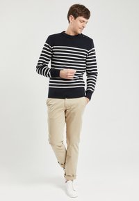 Maglione a righe blu e bianche con collo rotondo, dettaglio con bottoni sulla spalla, abbinato a pantaloni beige e sneakers bianche. Texture a maglia liscia.