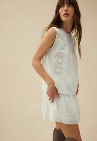 Robe sans manches blanche avec motifs floraux brodés et bordures en dentelle. Conception fluide avec une jupe froncée. Texture en coton lisse.