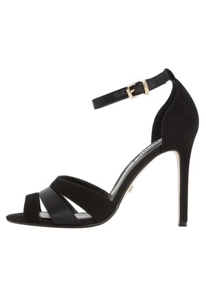 Sandal de tacón alto de ante negro con punta abierta, que presenta correas entrelazadas y una correa ajustable en el tobillo con herrajes en tono dorado.