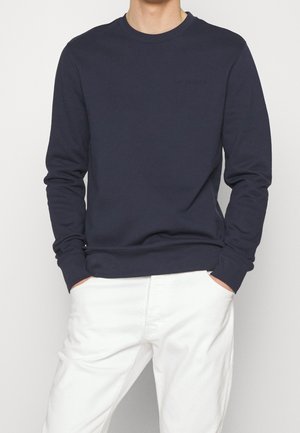 Man draagt een marineblauwe sweater met lange mouwen met handen in de zakken, gecombineerd met een witte broek tegen een effen achtergrond.