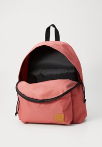 Mochila de tela rosa con interior negro, que cuenta con un bolsillo frontal, correas ajustables y un lazo para colgar. Cierre de cremallera y parche con logo.