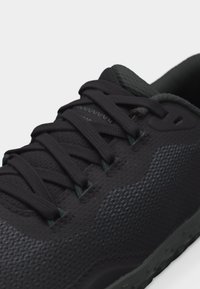 Κοντινή λήψη ενός μαύρου αθλητικού παπουτσιού με υφασμάτινη υφή, επένδυση στη γλώσσα και σφιχτά δεμένα μαύρα επίπεδα κορδόνια.