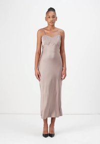 Abito in satin color taupe a sottoveste, con spalline sottili, un corpino aderente con dettagli arricciati e una gonna lunga fino al pavimento. Abbinato a tacchi neri.