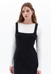 Schwarzes Denim-Kleid mit quadratischem Ausschnitt, dünnen Trägern und taillierter Silhouette. Getragen über einem weißen Langarmoberteil mit dezentem Muster.