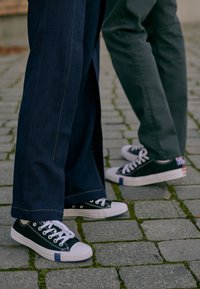 Baskets noires en toile avec des semelles en caoutchouc blanches et des accents bleus contrastants, associées à un pantalon évasé bleu foncé avec des coutures jaunes.