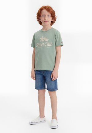 Dreng med rødt hår iført en grøn "Street Club" t-shirt, denimshorts og hvide slip-on sko foran en ensfarvet hvid baggrund.