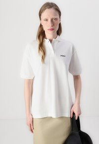 Biela bavlnená polo tričko s krátkymi rukávmi, so čiernym vyšitým logom na ľavej hrudi a uvoľneným strihom.