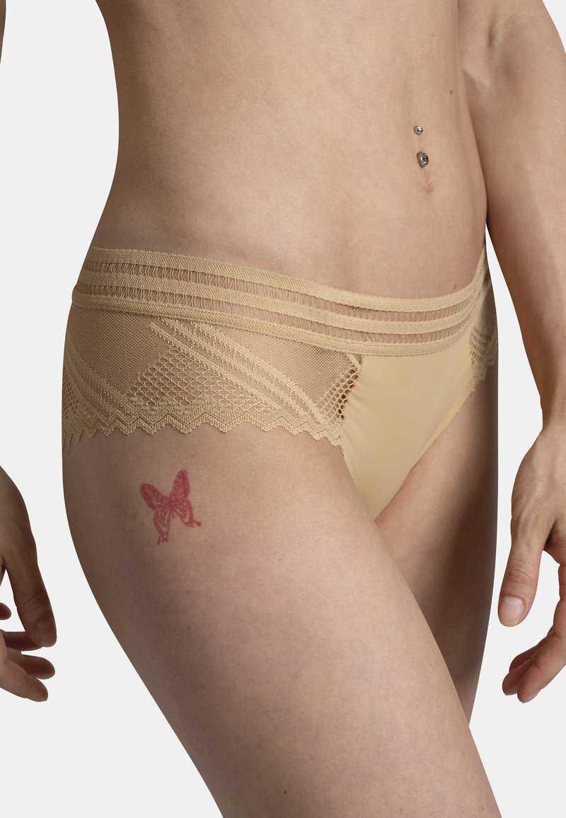 Sous-vêtement en dentelle beige avec une fine incrustation en maille et un tatouage papillon sur la cuisse, mettant en valeur la texture et le design.