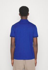 Modrá polo tričko s límečkem, krátkým rukávem a kontrastními pruhy na okrajích rukávů. Tkanina vypadá hladce a je mírně vypasované.