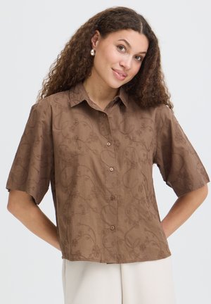 Femme aux cheveux bruns bouclés portant une chemise marron à manches courtes avec une broderie florale subtile, posant les mains dans le dos.