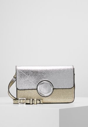 Mała metaliczna srebrna i złota torebka na ramię z okrągłym, przednim zapięciem i regulowanym paskiem, na białej powierzchni.