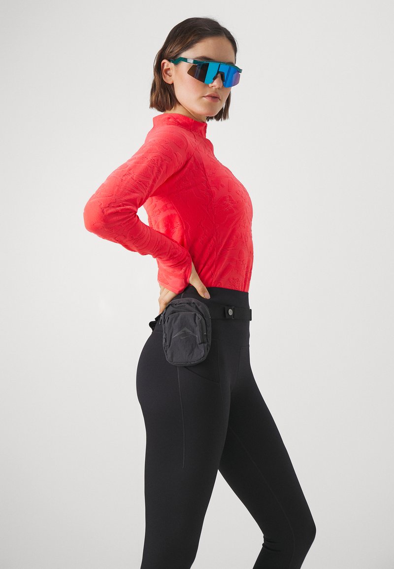 Haut long à manches longues texturé rouge associé à des leggings noirs, avec une poche latérale. Le modèle porte des lunettes de soleil bleues, se tenant de profil.