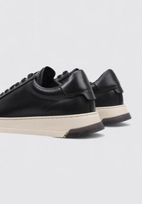Zapatillas bajas de cuero negro con parte trasera redondeada, suela beige en contraste y diseño con cordones que muestra una textura suave y detalles sutiles de costura.