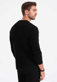 Suéter de punto negro con mangas largas, cuello redondo y superficie texturizada. Se lleva sobre unos pantalones vaqueros negros, mostrando una silueta ajustada por la parte de atrás.