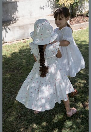 Δύο νεαρά κορίτσια με λευκά φορέματα παίζουν μαζί στο γρασίδι· η μία φοράει καπέλο με λουλούδια και έχει μακρύ κοτσίδα πλεξούδα.