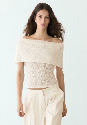 Frau mit langen, gewellten Haaren, die ein cremefarbenes Spitzen-Top mit freiliegenden Schultern und eine hoch taillierte beige Hose trägt, stehend vor einem einfarbigen hellen Hintergrund.