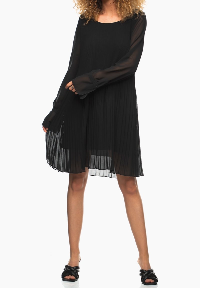 Femme en robe noire plissée à manches longues et sandales noires à bout ouvert, debout avec une main tenant l'ourlet de la robe.