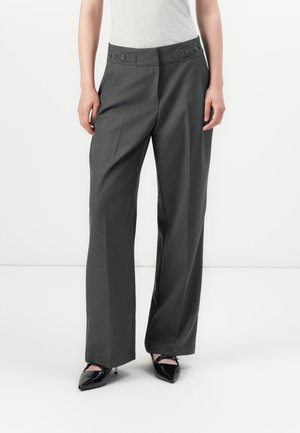 Person som bærer mørkegrå vide bukser med knappede midjbånd og svarte spisse hæler, står mot en hvit bakgrunn.