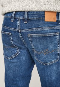 Vue arrière d'un jean en denim bleu avec des surpiqûres orange et une petite pièce en cuir marron sur la ceinture, porté avec un pull léger en maille.