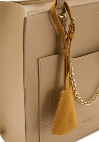 Beige leren handtas met een zachte textuur, voorzien van een gouden kettingaccent en een afneembare mosterdsuède label met reliëflogo.