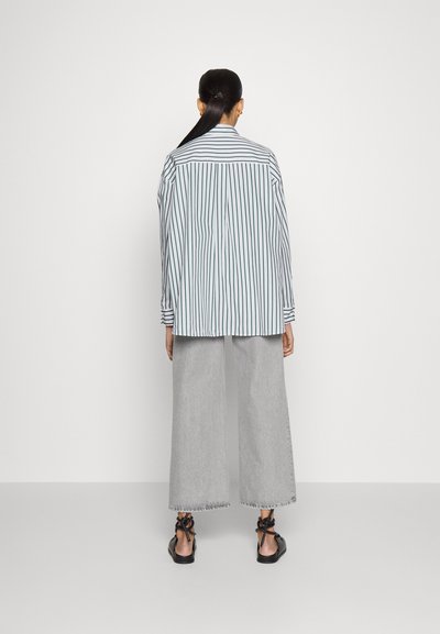 Camicia oversize a righe in azzurro chiaro e bianco, abbinata a jeans grigi a gamba larga e sandali neri con dettagli a fiocco.