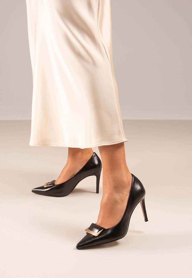Zapatos de tacón alto de cuero negro con puntera afilada y detalle de hebilla acentuado en oro. Presentados sobre un fondo claro, mostrando un diseño elegante.