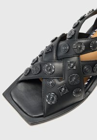 Open-toe black leather sandal with cross-strap design, embellished with decorative metal floral accents, contoured sole, and square toe.