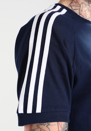 Navy blue athletic shirt with short sleeves, featuring white three-stripe accents on the shoulders and a crew neckline. Soft fabric texture.