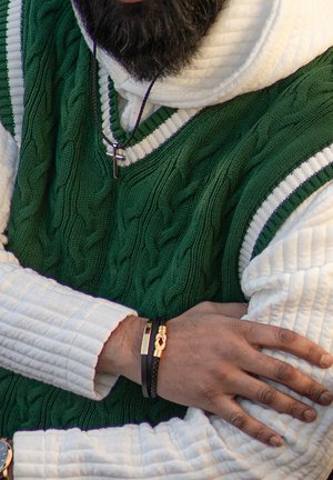 Green knitted vest with a cable pattern, layered over a white textured hoodie. Features black and gold bracelets on a partially visible arm.