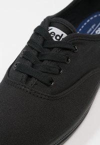 Musta kangaskengän tossu, jossa on teksturoitu pinta. Pyöreät kengännauhat ja metalliset nyörireiät. Kielen kohdalla valkoinen merkitty nauha. Kumipohja.