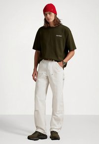 Young person wearing a red beanie, dark green oversized t-shirt, white loose pants, and olive green sneakers, standing with one hand in pocket.