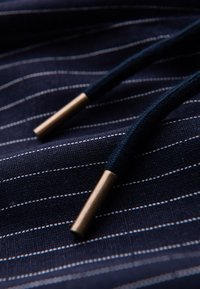 Gros plan d'un cordon noir avec embouts métalliques dorés sur un tissu bleu foncé à fines rayures blanches.