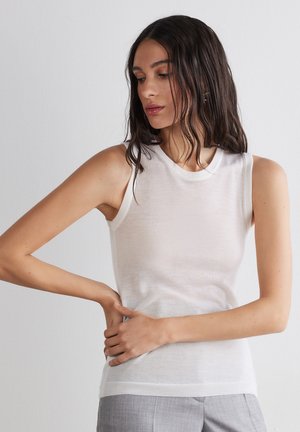 Femme aux longs cheveux foncés, vêtue d'un haut blanc sans manches et d'un pantalon gris, posant avec une main sur la hanche et regardant vers le bas.