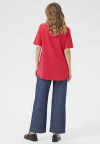 T-shirt in cotone rosso con maniche corte, orlo curvo, abbinato a pantaloni in denim a gamba larga e mocassini neri. Design semplice con colori solidi.