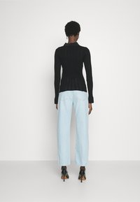 Persona con cabello corto y rizado, vistiendo una camiseta negra ajustada de manga larga, jeans rectos de mezclilla azul claro y zapatos de tacón alto negros, de pie de espaldas.