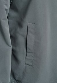 Šedá látková bunda s hladkou textúrou a bočným náprsníkom. Dizajn je minimalistický, s čistými líniami a bez ďalších vzorov.