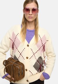 Kremowy kardigan z różowym i szarym wzorem argyle, brązowe guziki, zestawiony z lawendowym swetrem, dużymi różowymi okularami przeciwsłonecznymi i brązową torbą.