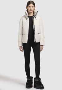 Veste blanche rembourrée avec un col haut, fermeture éclair à l'avant et détails en noir, portée sur un haut noir et des leggings ajustés avec des bottes noires.