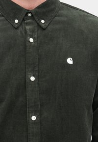 Παντελόνι από βελούδινο ύφασμα σε σκούρο πράσινο χρώμα, με γιακά που κλείνει με κουμπιά, λευκά κουμπιά και μικρό λογότυπο στη αριστερή πλευρά. Υφή υφάσματος με κάθετες ρίγες.