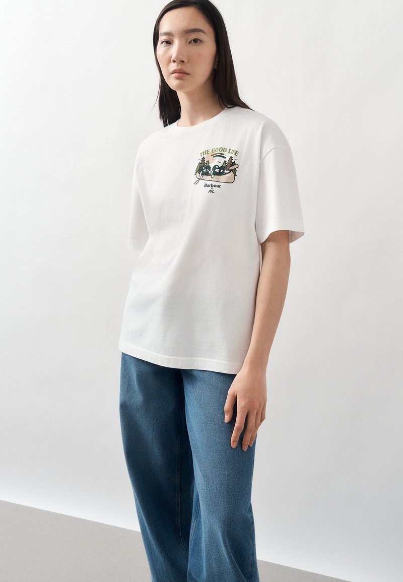 Γυναίκα που φοράει λευκό T-shirt με γραφικό ρακούν, το κείμενο «The Good Life» και μπλε τζιν, στέκεται σε απλό φωτεινό φόντο.